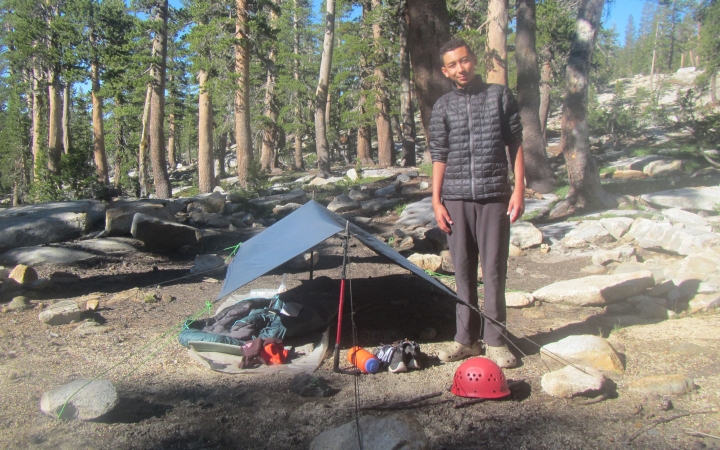 a person stands beside their tarp shelter in a wooded area on an outward bound trip